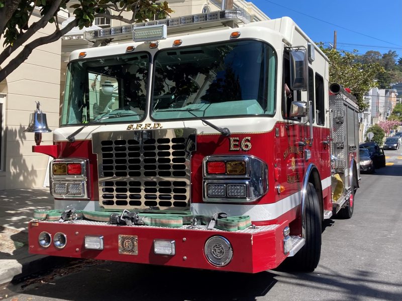 A red and white fire truck with "S.F.F.D." and "E6" markings is parked on a street next to a light-colored building. Trees and a clear blue sky are in the background.