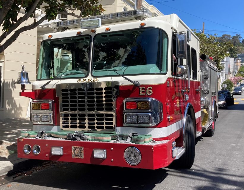 A red and white fire truck with "S.F.F.D." and "E6" markings is parked on a street next to a light-colored building. Trees and a clear blue sky are in the background.
