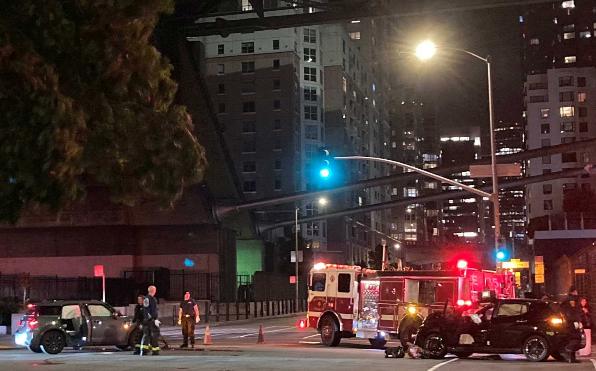 Nighttime roadside scene with emergency response: two damaged cars, a firetruck with lights on, and several responders on the road under city streetlights with tall buildings in the background.