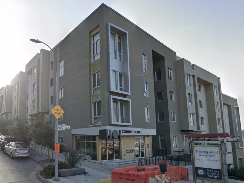 A modern multi-story apartment building with a store on the ground floor. Construction barriers and a street intersection are visible in the foreground.