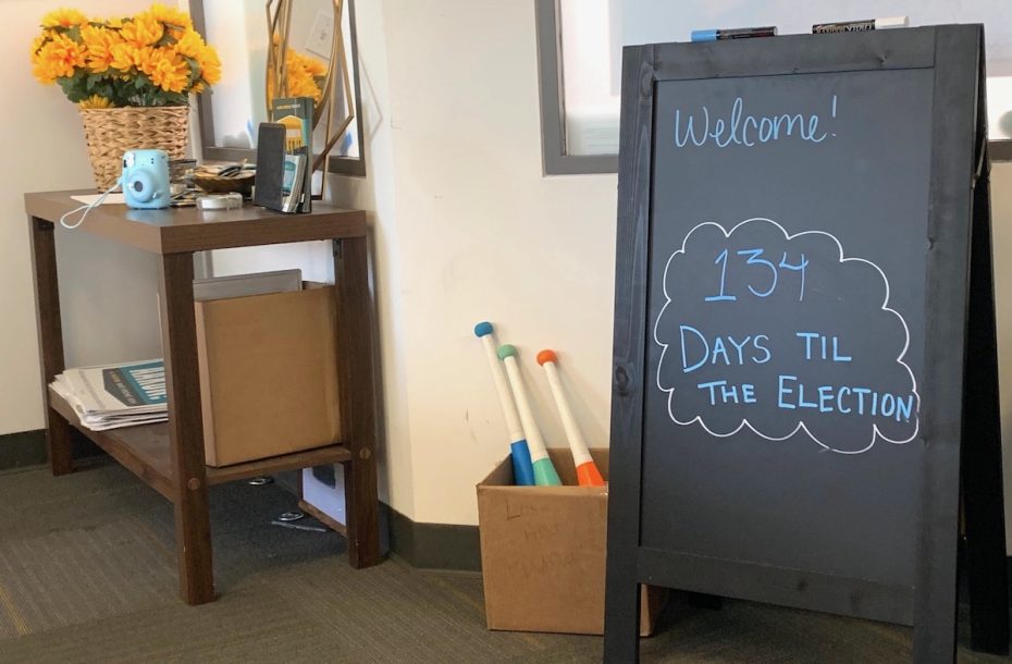 A chalkboard sign in an office reads "Welcome! 134 days til the election." Nearby, a table holds a vase with sunflowers and various items, and a box on the floor contains traffic cones.