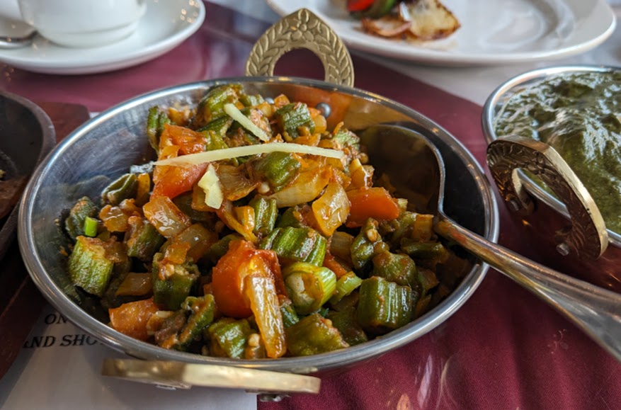 A stainless steel bowl filled with a vegetable curry, including okra, tomatoes, and onions, is placed on a table next to a cup, a plate, and another dish.