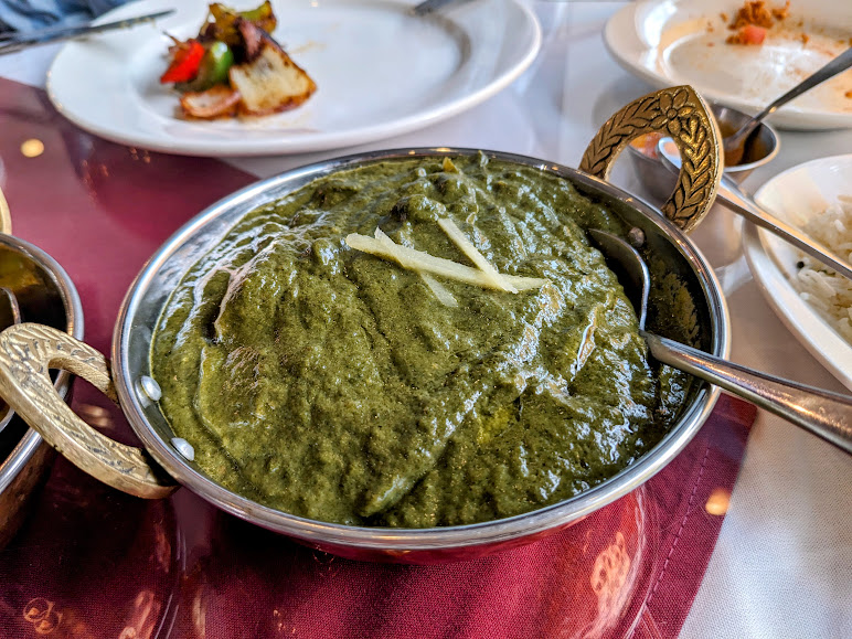A bowl of green creamy Indian curry garnished with ginger strips. A spoon is placed in the curry. Other food items and plates are visible in the background.