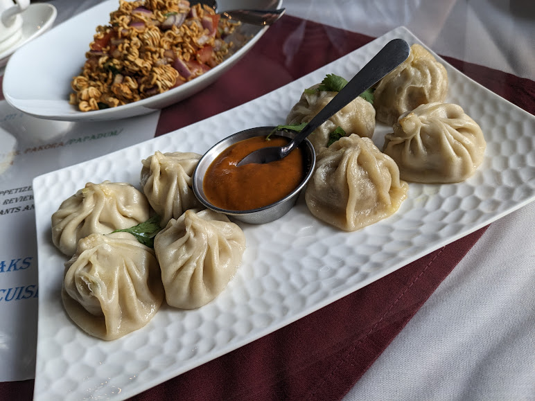 Plate of dumplings served with a small bowl of orange dipping sauce and a spoon, alongside another dish topped with crispy noodles in the background.