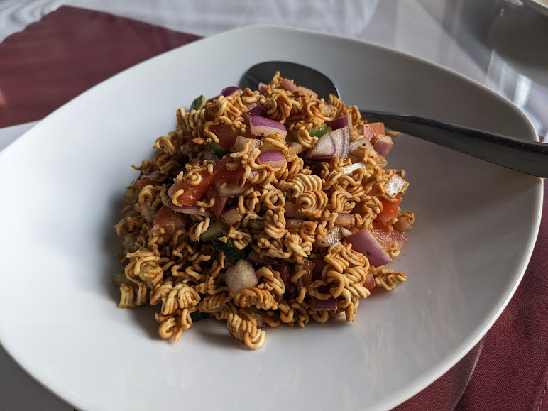 A white plate with a serving of dry, seasoned instant noodles mixed with chopped vegetables and red onion, accompanied by a metal spoon.