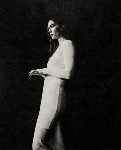 Black and white image of a woman standing against a dark background, wearing a long-sleeved dress and looking to the left with hands clasped.