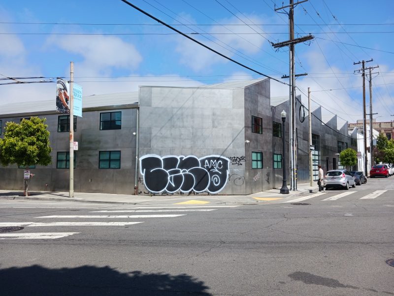 A street corner featuring a gray building with the word "Ouch" painted on the side, power lines above, and a few parked cars.