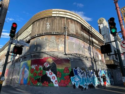 A weathered building is partially covered with vibrant wall art and graffiti, located at the intersection of Mission Street. Traffic lights and street signs are visible under a clear blue sky.