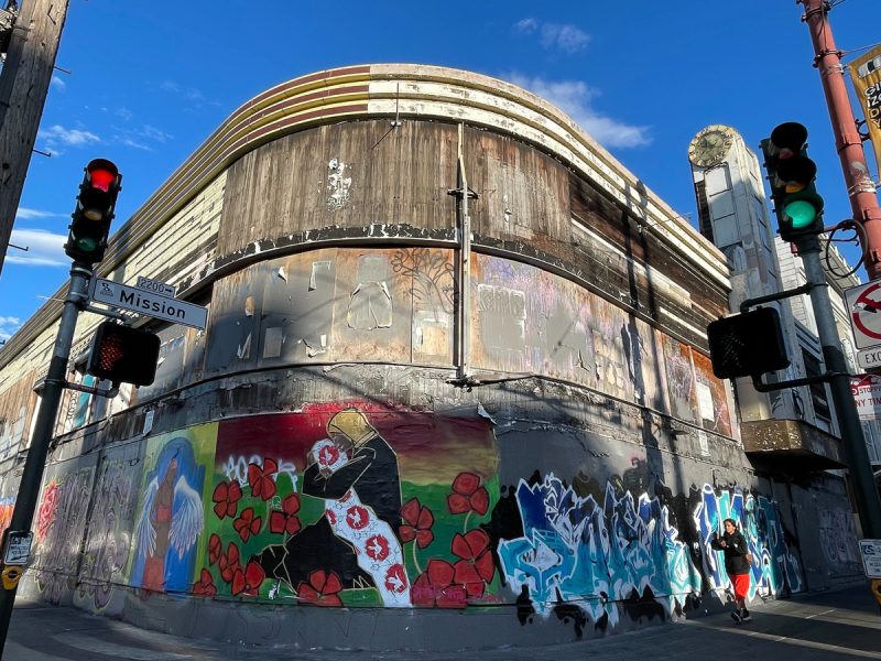A weathered building is partially covered with vibrant wall art and graffiti, located at the intersection of Mission Street. Traffic lights and street signs are visible under a clear blue sky.