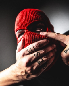 A person wearing a red knit balaclava covers their mouth with tattooed hands.