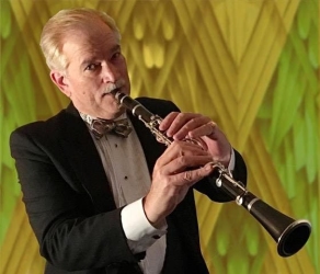 A man in a tuxedo plays a clarinet in front of a patterned gold and green background.