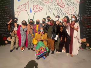 A group of people, wearing a mix of colorful and neutral outfits, pose for a photo in front of a floral-themed backdrop. Most of them are wearing face masks.