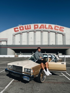 A person sits on the hood of a beige car with an open door in an empty parking lot, in front of a building labeled "Cow Palace.