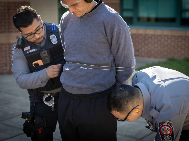 Two police officers are securing a man with zip ties. The man is wearing a gray sweatshirt. This interaction is taking place outdoors, in front of a brick building.