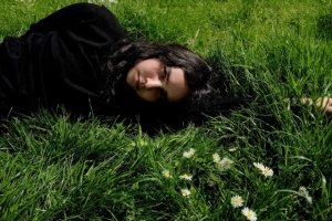 A person with long dark hair lying on green grass with daisies nearby, wearing a black outfit, looking at the camera.