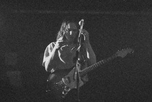 Black and white image of a person standing on stage, holding a guitar and adjusting a microphone.