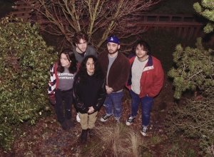 Five people stand together outdoors surrounded by greenery, looking up at the camera. They wear casual clothing and some have jackets on. The background includes trees and shrubs.
