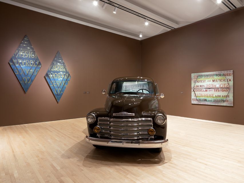 A vintage Chevrolet truck is showcased in a museum-like art gallery, its vibrant presence complementing paintings on brown walls and wood flooring.
