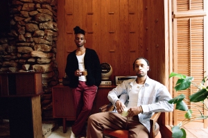 Two individuals are in a room with wood-paneled walls and a stone wall. One stands holding a mug, and the other sits with a cup. There is green plant foliage in the foreground.