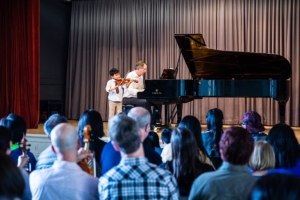 A young child plays the violin next to an adult pianist on stage while an audience watches in a concert hall.