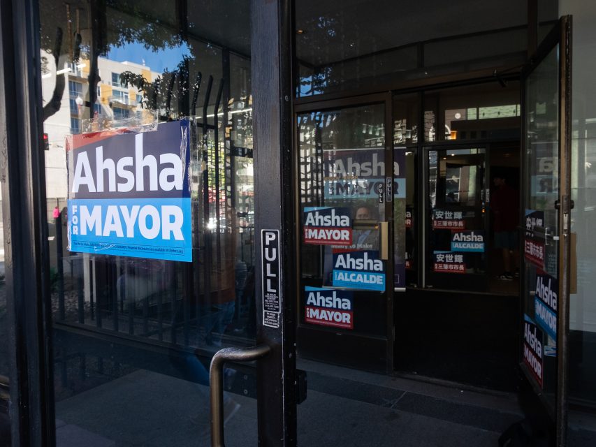 Glass door with "Ahsha for Mayor" campaign signs and multiple similar signs visible inside the building. The door has a 'PULL' label. Trees and residential buildings are reflected in the glass.
