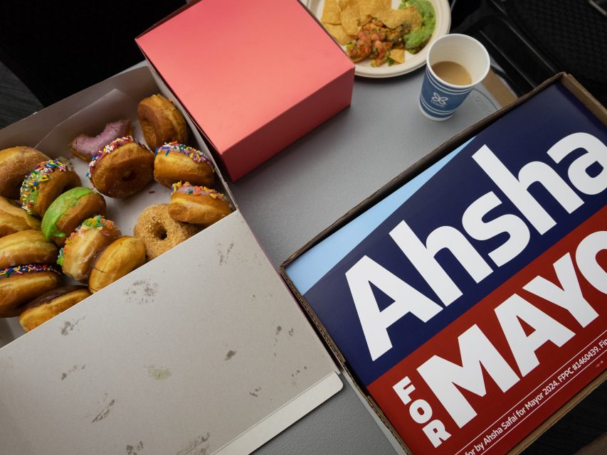 Open boxes with donuts and a "Ahsha for Mayor" campaign sign on a table, with a cup of coffee and a plate of food in the background.