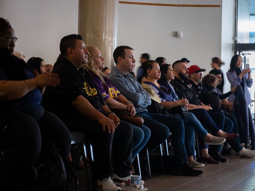 A group of people sits attentively in rows of chairs, facing forward in a room with neutral-colored walls and a large window to the right.