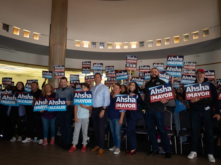 A group of people in a room holds signs supporting a mayoral candidate named Ahsha. They are posing and smiling in front of a circular room with a high ceiling and windows.