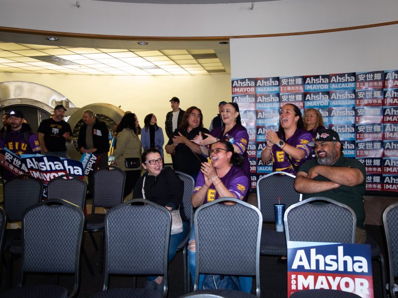 A group of seated people, some standing and clapping, are gathered in a room with campaign signs reading "Ahsha for Mayor" on the walls.