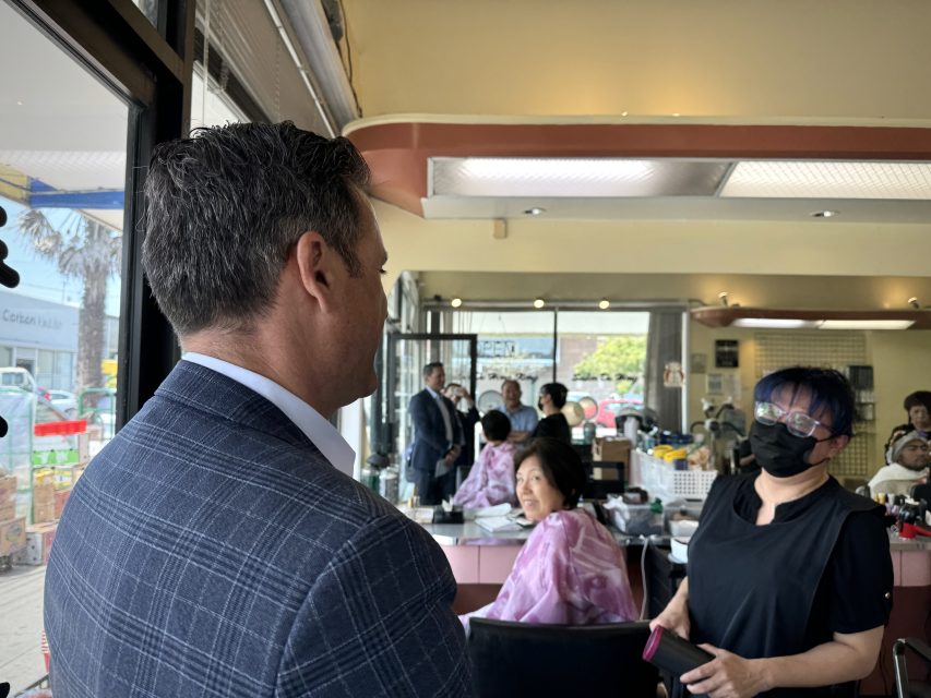 在一家繁忙的美髮沙龍裡，一名身着格子西裝外套的男子與一名戴着口罩的黑衣女子交談，沙龍裡有幾名顧客和髮型師。