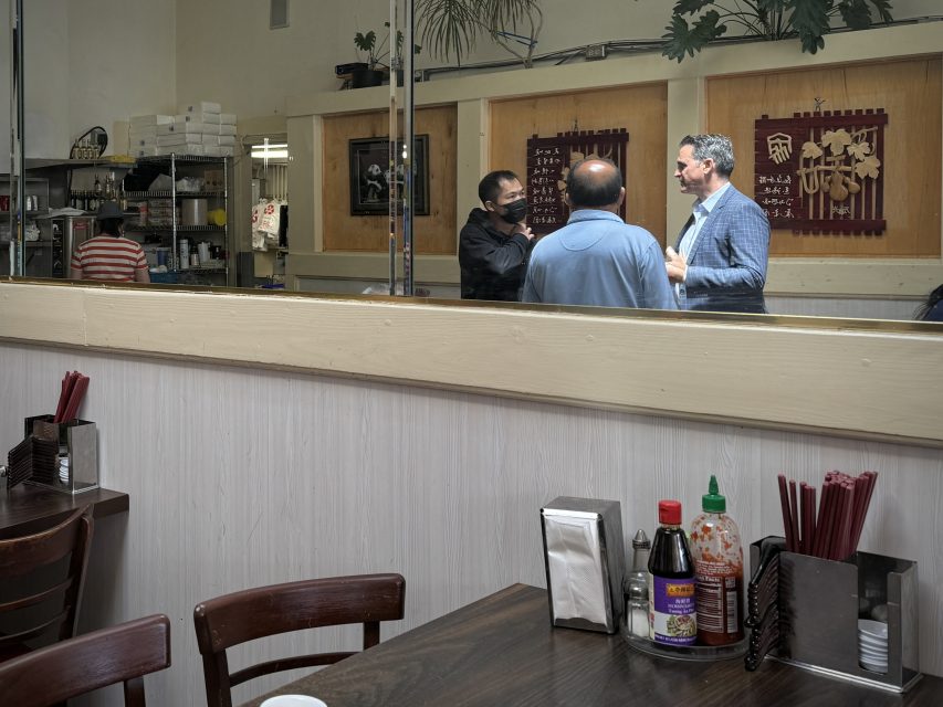 三名男子在一家餐廳內交談，背景是廚房。前景的桌子上擺放着調味品和筷子。