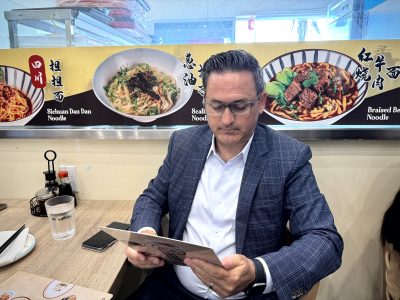 A man in a blue plaid suit jacket and white shirt reads a menu while seated in a restaurant. Behind him are images of Asian noodle dishes with descriptions. A glass of water and condiments are on the table.