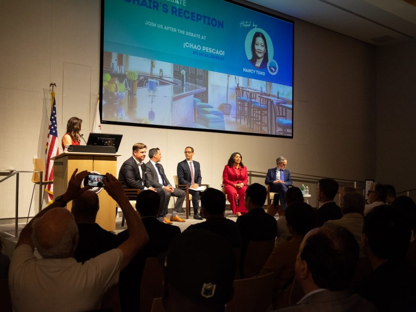 A panel of five people is seated on a stage in front of a seated audience. A woman stands at a podium speaking. An American flag is visible.