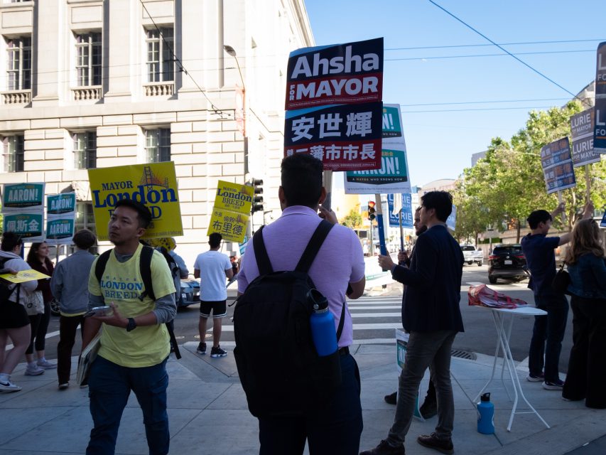 A group of people standing on a city street hold signs promoting various mayoral candidates.