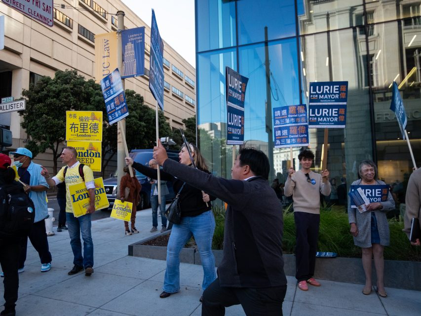 Group of people on a city sidewalk holding signs supporting two different mayoral candidates. Buildings and offices are in the background.