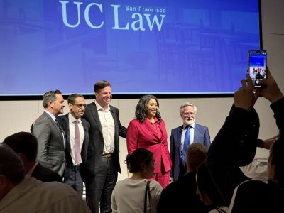 Five individuals are standing in front of a screen displaying "UC Law San Francisco." One person in the crowd is capturing the moment with a phone.