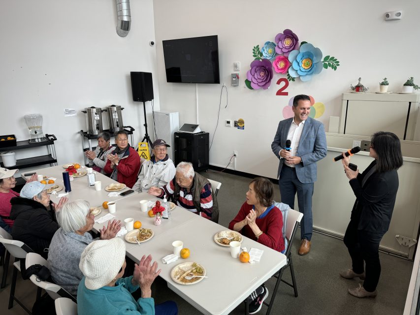 人们端着餐盘坐在桌边，旁边站着两个人，手里拿着麦克风。房间的墙上挂着装饰品，包括五颜六色的花朵和一个大电视屏幕。