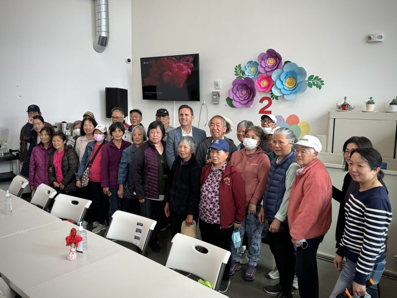 A group of people, including some elderly individuals, pose together for a photo in a room decorated with colorful paper flowers. A man in a suit stands in the center of the group.