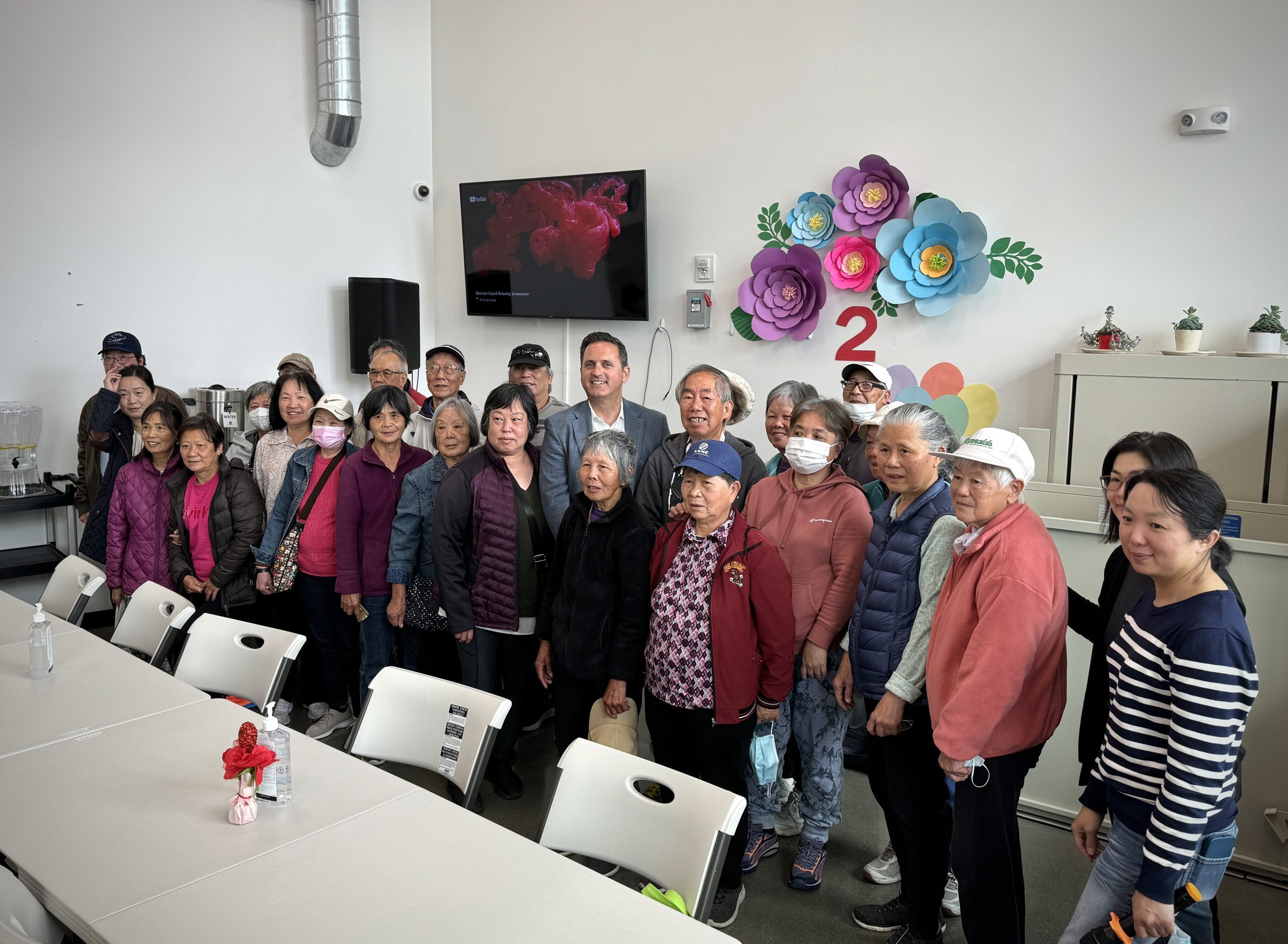 A group of people, including some elderly individuals, pose together for a photo in a room decorated with colorful paper flowers. A man in a suit stands in the center of the group.