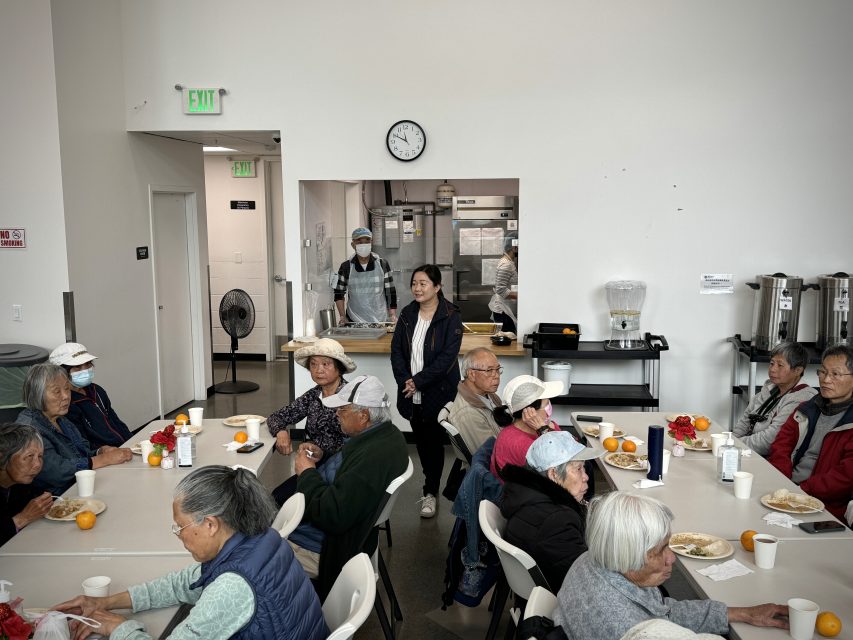 一群老年人坐在自助餐厅的桌边交谈。一些人正在用餐，背景是厨房里的工人。