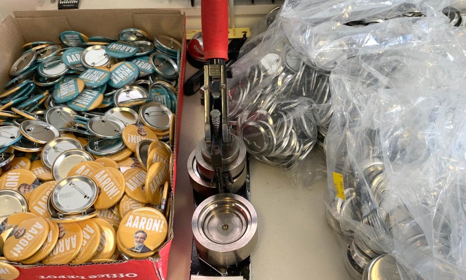 A table with a button-making machine, a box of completed buttons labeled "AARON!" with a picture of a person, and plastic bags filled with metal button parts.