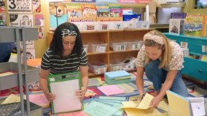 Two women organize educational materials on the floor in a colorful classroom filled with books and posters.
