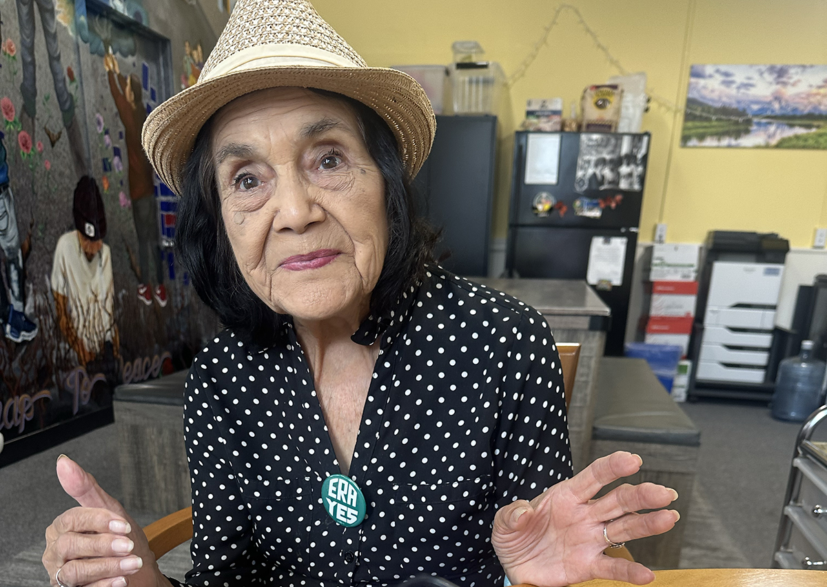 An older woman with dark hair wearing a straw hat and a black polka dot blouse is sitting indoors and speaking. She has a badge on her blouse that reads "ERA YES.