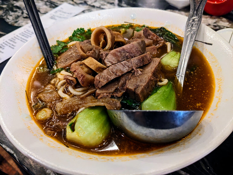 A bowl of beef noodle soup with bok choy, cilantro, and sliced beef, served with a large soup spoon and chopsticks.