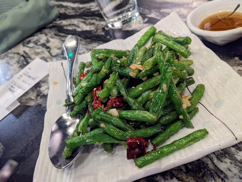 A plate of stir-fried green beans with garlic and red chili peppers, served with a spoon on the side. A glass of water and a small bowl with dipping sauce are in the background.
