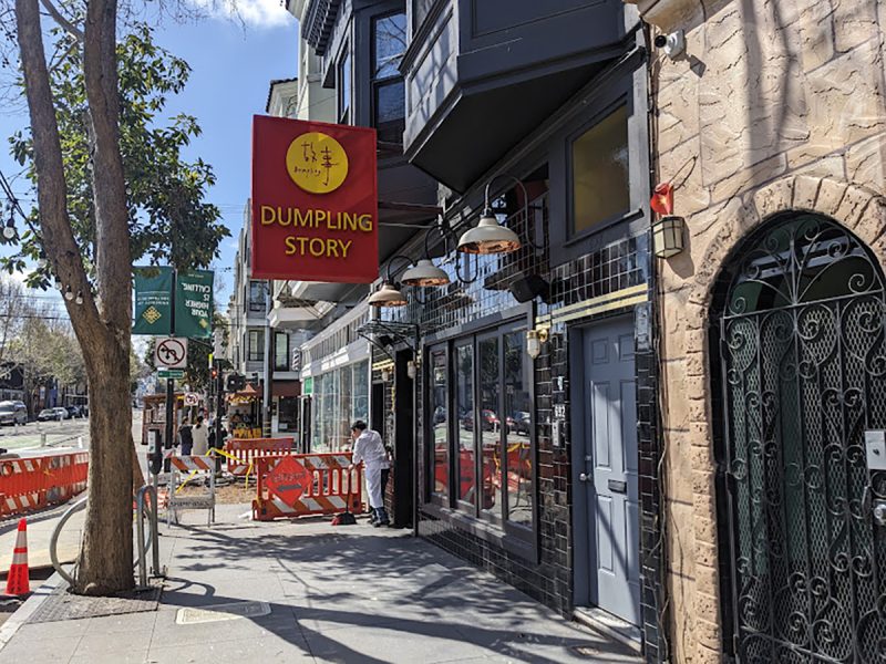 A sidewalk view of a restaurant named "Dumpling Story" with a red sign and Chinese characters. An individual is performing maintenance near an orange barrier outside the entrance.