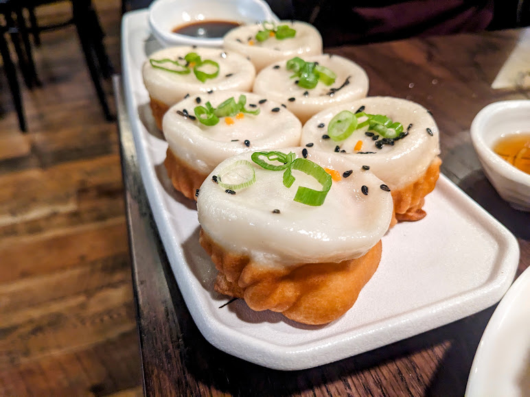 A plate of six golden-brown Chinese dumplings topped with green onions and black sesame seeds, accompanied by a dish of soy sauce on the side.