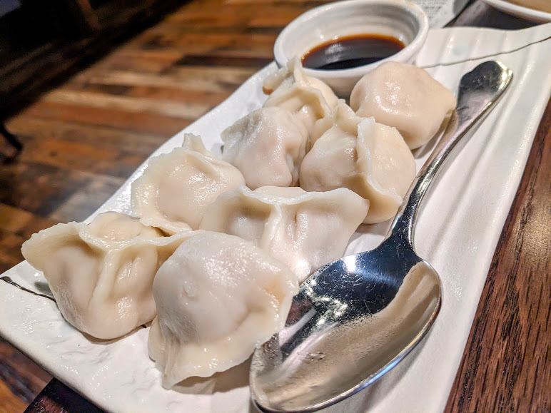 A white plate of dumplings with a silver spoon and a small bowl of dipping sauce on a wooden table.