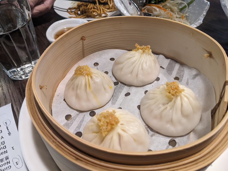 Four steamed dumplings in a bamboo steamer basket, garnished with crispy bits, with a glass of water, a bowl of sauce, and a plate of noodles in the background.