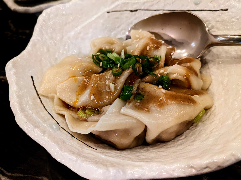 A white ceramic bowl with dumplings topped with chopped green onions, sesame seeds, and brown sauce, accompanied by a metal spoon.